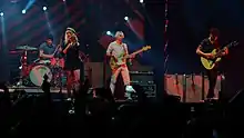 A band performs for a live audience. One man on guitar, one on drums, one on bass, and the frontwoman singing into a microphone.